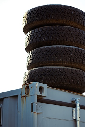A stack of four dark, rugged tires is placed on top of a weathered metal container with visible rust stains and exposed metal hinges.