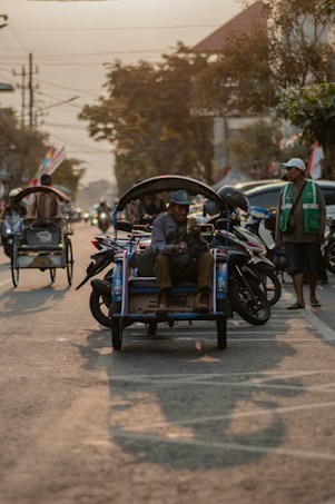 A street scene featuring a man sitting on a traditional tricycle taxi, known as a becak, while holding a mobile phone. Surrounding him are motorcycles and another man wearing a green vest, standing nearby. The background includes trees, some buildings, and vehicles with the warm glow of the setting sun casting long shadows on the road.