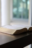 An open book resting on a wooden table by a window.