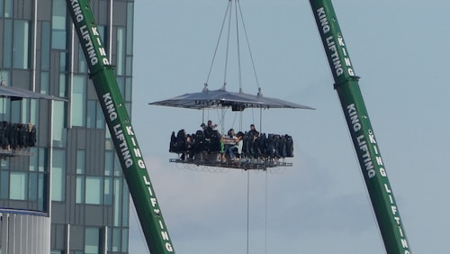 A platform suspended by cranes is elevated high above the ground, with people seated around what appears to be a dining table. The setup includes two green cranes labeled 'KING LIFTING' on either side. The background features a modern high-rise building, adding an urban context to the scene.