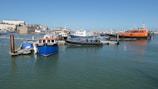 Several boats are docked at a harbor, with various types and sizes of vessels including a blue boat named Sea Searcher, a black inflatable boat, and an orange pilot boat. The harbor is calm with clear water reflecting the boats, and buildings are visible in the background under a clear blue sky.