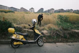 A yellow and black motor scooter with a white helmet on the seat is parked on the side of a road. In the background, a person is bent over working or examining something in the tall grass adjacent to the road. Behind the grassy area, there are rows of multi-story apartment buildings in the distance.