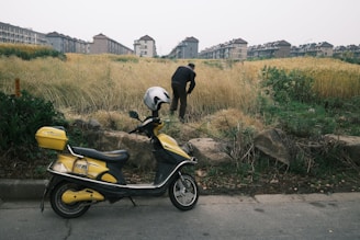 A yellow and black motor scooter with a white helmet on the seat is parked on the side of a road. In the background, a person is bent over working or examining something in the tall grass adjacent to the road. Behind the grassy area, there are rows of multi-story apartment buildings in the distance.