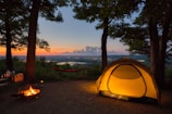 A serene campsite set in a wooded area at sunset, featuring a glowing tent illuminated from within. A hammock is strung between two trees, and a small campfire burns nearby. Beyond the campsite, the landscape stretches into the distance with a river or lake reflecting the evening light.