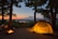 A serene campsite set in a wooded area at sunset, featuring a glowing tent illuminated from within. A hammock is strung between two trees, and a small campfire burns nearby. Beyond the campsite, the landscape stretches into the distance with a river or lake reflecting the evening light.