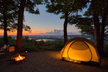 A serene campsite set in a wooded area at sunset, featuring a glowing tent illuminated from within. A hammock is strung between two trees, and a small campfire burns nearby. Beyond the campsite, the landscape stretches into the distance with a river or lake reflecting the evening light.