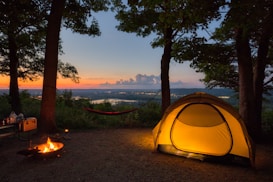 A serene campsite set in a wooded area at sunset, featuring a glowing tent illuminated from within. A hammock is strung between two trees, and a small campfire burns nearby. Beyond the campsite, the landscape stretches into the distance with a river or lake reflecting the evening light.