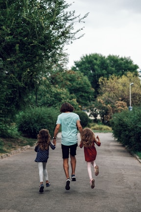A man and two children, all holding hands, walk down a path surrounded by greenery. The children have long hair and wear colorful clothes, while the man is dressed casually in a light blue shirt and shorts. The scene captures a moment of togetherness and nature.