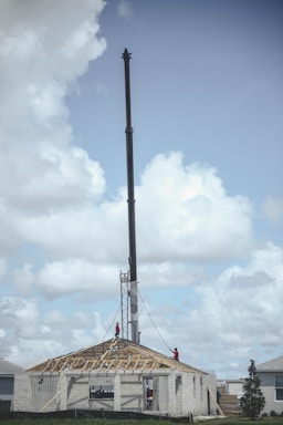A construction site with a crane towering over a building under construction. Workers in red safety gear are working on the roof framing of the structure. The sky is partly cloudy with soft, white clouds.