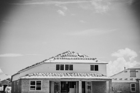 A house under construction with roofing materials stacked on top. The building appears to be in the framing stage with partially completed windows and exterior walls. Other partially built houses are visible in the background under a partly cloudy sky.