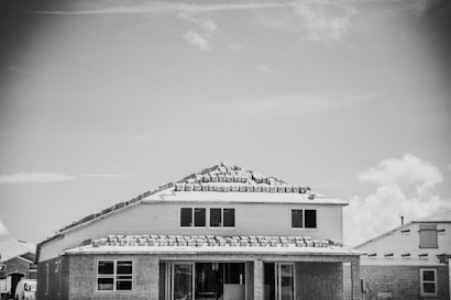 A house under construction with roofing materials stacked on top. The building appears to be in the framing stage with partially completed windows and exterior walls. Other partially built houses are visible in the background under a partly cloudy sky.