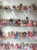 A collection of dolls displayed on shelves, each wearing an eye mask. The dolls are dressed in a variety of outfits ranging from casual to formal. Their hairstyles vary, and they are positioned sitting or leaning against the back of the shelves.