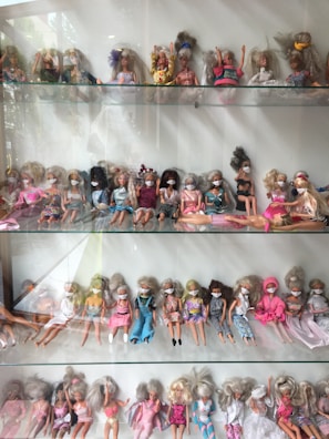 A collection of dolls displayed on shelves, each wearing an eye mask. The dolls are dressed in a variety of outfits ranging from casual to formal. Their hairstyles vary, and they are positioned sitting or leaning against the back of the shelves.