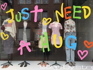 A storefront window display featuring five mannequins dressed in various casual outfits. The window is decorated with large, colorful letters spelling 'JUST NEED A HUG' along with heart shapes. The outfits include a striped sweater with an American flag, a plaid shirt, a pink checkered dress, a floral blouse with ripped jeans, and other trendy garments. The display conveys a warm and inviting message.