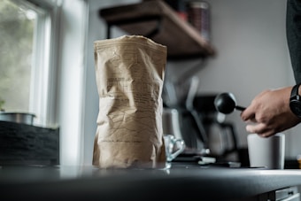 A close-up of a paper coffee bag labeled 'Ethiopia' is placed on a kitchen countertop near a window. Next to it, a person holding a coffee scoop stands, partially visible, suggesting coffee preparation. In the blurred background, various kitchen items and utensils create a cozy atmosphere.