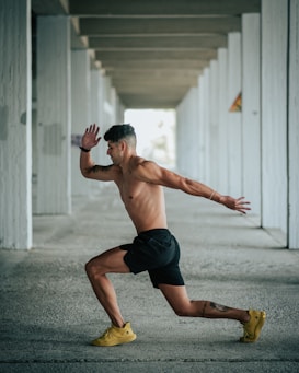 A shirtless man in black shorts and yellow shoes is captured in a dynamic pose, appearing to be in motion as if running or lunging forward. The setting is an architectural structure with repetitive vertical columns and a concrete floor, creating a sense of depth and perspective.