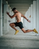 A shirtless man is captured in mid-air while performing a leap inside a concrete structure. He is wearing black shorts and yellow athletic shoes and has a fit physique. The background comprises multiple gray concrete pillars forming a repetitive pattern.