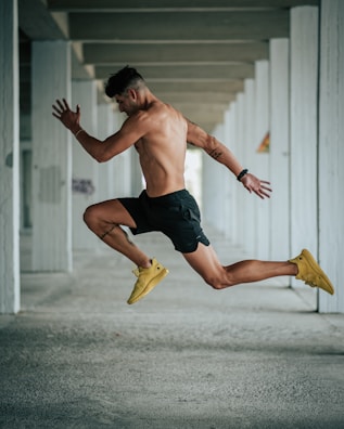 A shirtless man is captured in mid-air while performing a leap inside a concrete structure. He is wearing black shorts and yellow athletic shoes and has a fit physique. The background comprises multiple gray concrete pillars forming a repetitive pattern.