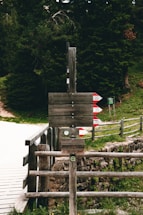 A rustic wooden signpost in the forest pointing toward the camp with handcarts and tents visible in the background.
