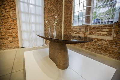 A sleek, modern table with a curved wooden base is placed on a white platform. Three delicate, minimalist vases with white flowers decorate the tabletop. The background consists of exposed brick walls, large windows, and white sheer curtains, creating an urban yet elegant atmosphere.