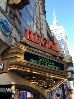 A movie theater marquee with a prominent sign displaying the words 'REGAL CINEMAS' in red above an LED display advertising the movie 'GODZILLA KING OF THE MONSTERS'. The building features ornate architectural details and is surrounded by various other signs and buildings in an urban setting.