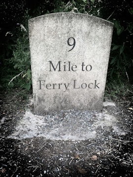 A weathered stone milestone marker with '9 Mile to Ferry Lock' etched on its surface, surrounded by grass and foliage.