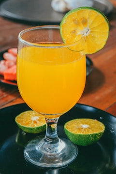 A glass filled with vibrant orange juice is garnished with a slice of lime on the rim. The glass stands on a dark plate with two halves of a lime beside it. The background features a wooden surface, adding a rustic touch to the scene.