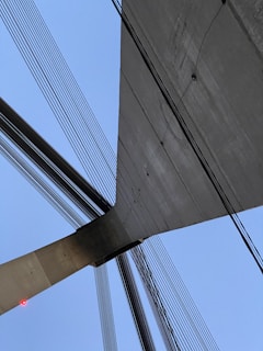A close-up view of a modern suspension bridge with numerous cables extending in various directions. The structure is composed of concrete and steel, emphasizing its sleek, architectural design. The sky forms a clear blue background, enhancing the contrast with the bridge's materials. A small red light is visible on one end of the concrete structure.