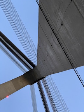 A close-up view of a modern suspension bridge with numerous cables extending in various directions. The structure is composed of concrete and steel, emphasizing its sleek, architectural design. The sky forms a clear blue background, enhancing the contrast with the bridge's materials. A small red light is visible on one end of the concrete structure.