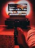 A hand holds a remote with visible buttons, pointed towards a wall-mounted TV displaying a streaming platform interface. The room is dimly lit, with a warm, red ambient light highlighting the TV screen and surroundings.