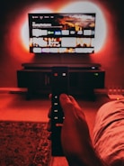 A hand holds a remote with visible buttons, pointed towards a wall-mounted TV displaying a streaming platform interface. The room is dimly lit, with a warm, red ambient light highlighting the TV screen and surroundings.
