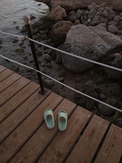 A pair of pale green slippers rests on a wooden deck that overlooks a rocky shoreline. The deck is secured with a black and white railing, and the background features large, weathered rocks leading down to the water.