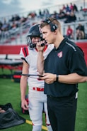 A coach is holding a clipboard and wearing a headset while talking to a player who is dressed in a football uniform. They are standing on a synthetic turf field with bleachers and spectators in the background.