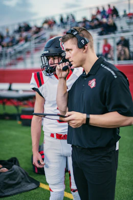 A coach is holding a clipboard and wearing a headset while talking to a player who is dressed in a football uniform. They are standing on a synthetic turf field with bleachers and spectators in the background.