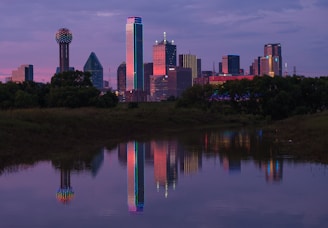 a city skyline is reflected in the still water of a pond