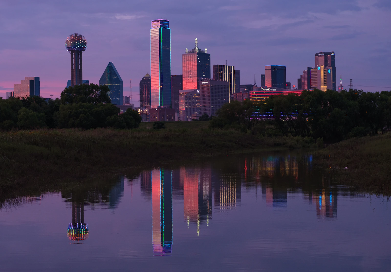 Dallas skyline reflected in calm water at dusk with a restrained editorial color palette.