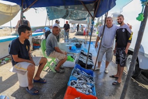 A bustling outdoor fish market with several men engaging in conversation and selling fish. Blue crates filled with various fish are displayed on the ground. The scene is set on a sunny day near the coast, with some boats visible in the background.
