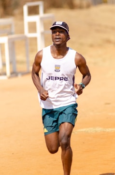 A person is running on a dirt track, wearing a white tank top with a logo and green shorts. The setting appears to be outdoors in a bright, sunny environment with a blurred background.