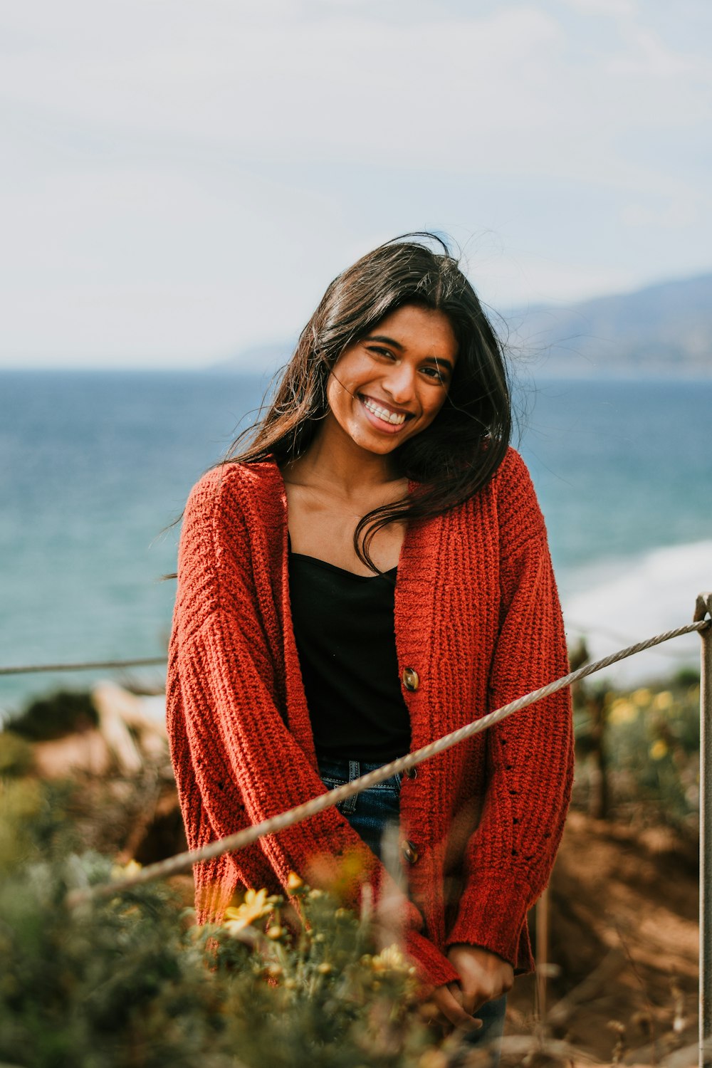 a smiling woman in a red cardigan by the ocean