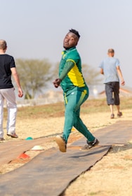 A person in a green and yellow sports uniform is in mid-motion, likely participating in a sporting event on a grassy field. Two other individuals are walking away in the background. The setting appears outdoor with clear skies and scattered trees.