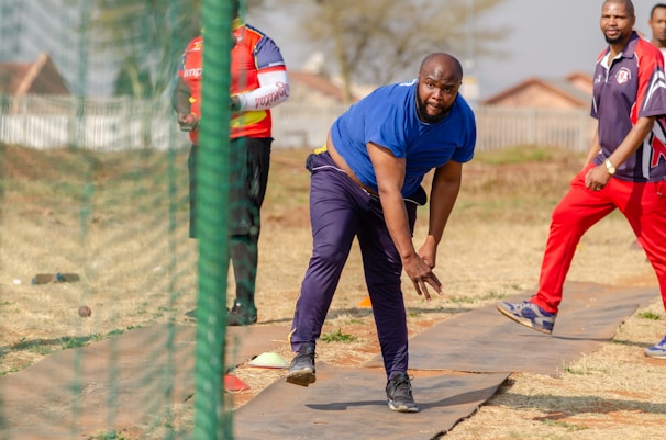 A man in a blue shirt and dark pants is bowling on a cricket pitch, while two other men, one wearing a bright sports jersey and the other in a dark sports outfit, watch him. The setting appears to be an outdoor area with dry grass and a net surrounding the playing area.
