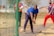 A man in a blue shirt and dark pants is bowling on a cricket pitch, while two other men, one wearing a bright sports jersey and the other in a dark sports outfit, watch him. The setting appears to be an outdoor area with dry grass and a net surrounding the playing area.