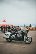 A stylish motorcycle is parked in an open lot with a custom paint design featuring flames on its body. In the background, a large red and white tent is set up, and several people gather around, creating a lively atmosphere reminiscent of a fair or gathering. The sky is overcast, suggesting a cloudy day.