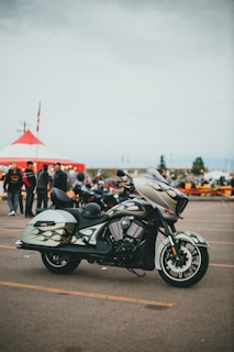 A stylish motorcycle is parked in an open lot with a custom paint design featuring flames on its body. In the background, a large red and white tent is set up, and several people gather around, creating a lively atmosphere reminiscent of a fair or gathering. The sky is overcast, suggesting a cloudy day.