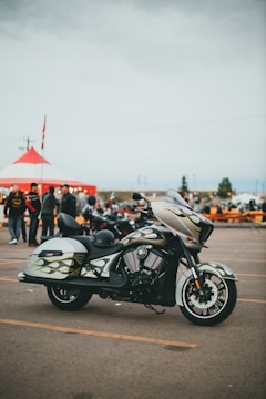 A stylish motorcycle is parked in an open lot with a custom paint design featuring flames on its body. In the background, a large red and white tent is set up, and several people gather around, creating a lively atmosphere reminiscent of a fair or gathering. The sky is overcast, suggesting a cloudy day.