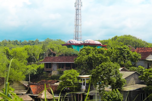 A rural setting with several houses featuring red-tiled roofs surrounded by lush greenery and trees. In the background, a large metal tower rises above the landscape, and a colorful, dome-shaped structure with a teal base and red trim is visible among the trees.