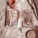 A cozy tabletop scene with an open journal displaying a decorative page filled with handwritten text, illustration cutouts, and scrapbook-style elements. Beside the journal, a lit pink candle adds warmth and ambiance to the setting. The background consists of a soft fabric with a textured appearance, and there is a round wooden slice in the corner of the image.