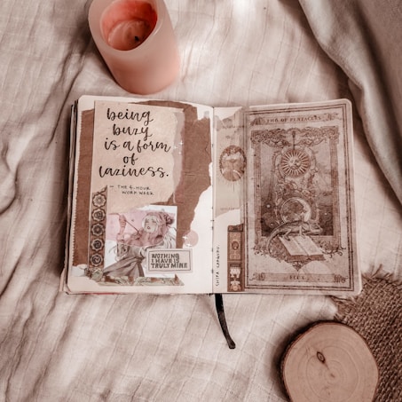 A cozy tabletop scene with an open journal displaying a decorative page filled with handwritten text, illustration cutouts, and scrapbook-style elements. Beside the journal, a lit pink candle adds warmth and ambiance to the setting. The background consists of a soft fabric with a textured appearance, and there is a round wooden slice in the corner of the image.