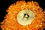 A golden Bitcoin coin is prominently placed among a pile of shiny, small orange beads. The background is black, highlighting the contrast between the glowing objects and their surroundings. The design on the Bitcoin is intricate and distinct.