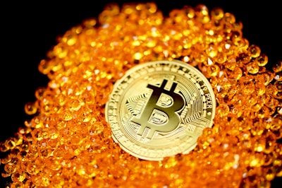 A golden Bitcoin coin is prominently placed among a pile of shiny, small orange beads. The background is black, highlighting the contrast between the glowing objects and their surroundings. The design on the Bitcoin is intricate and distinct.